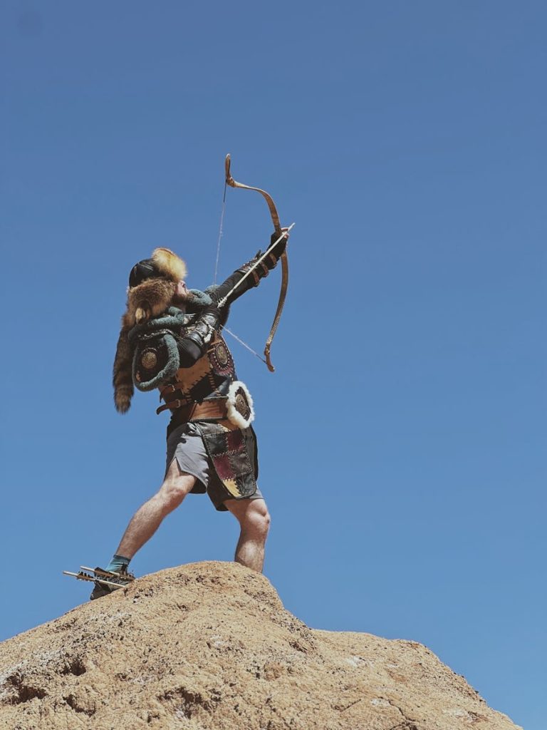 An archer in armor aims a bow towards the sky on a sunny day in a desert terrain.