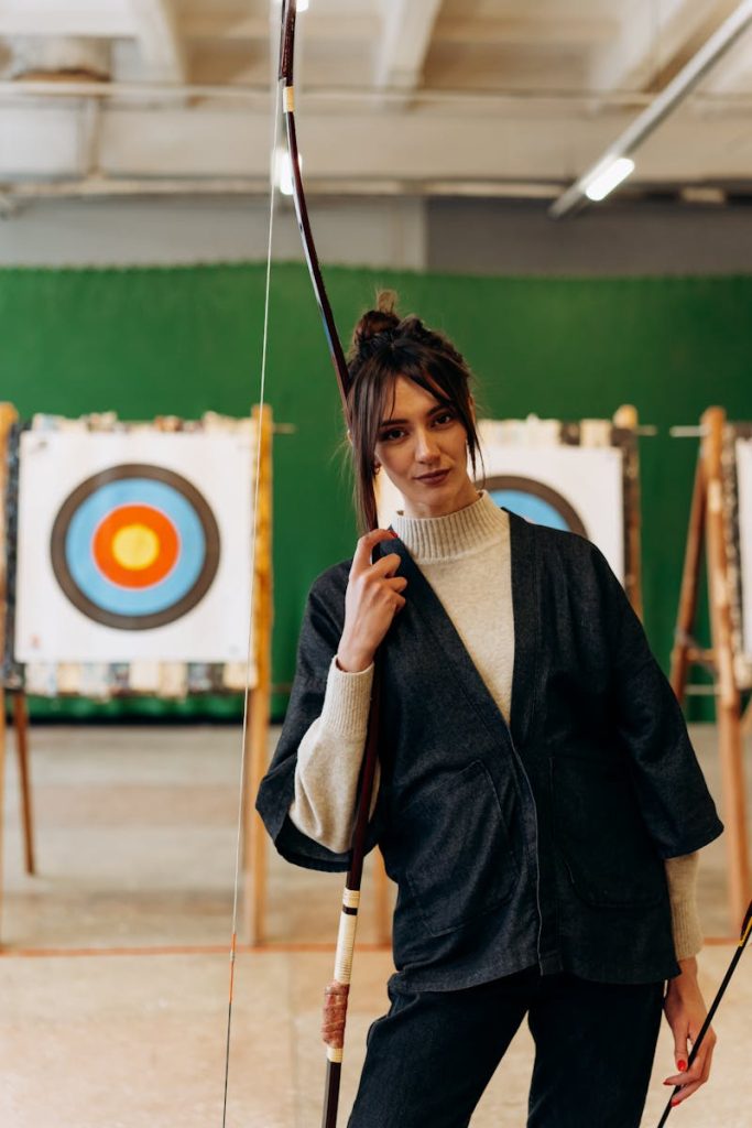 Confident young woman poses with bow in an indoor archery range, embodying focus and strength.