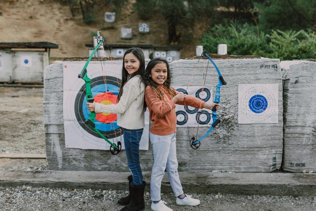 Two young girls practicing archery outdoors with target boards and bows, embracing leisure and sport.